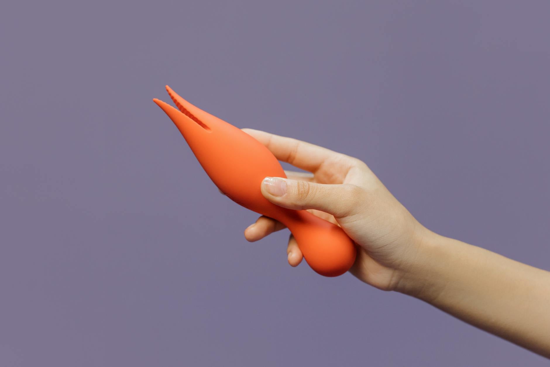 A hand holding a bright orange vibrator against a minimalistic purple backdrop